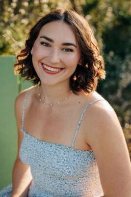 Headshot of Alaina Neuburger. Neuburger has shoulder-length wavy brown hear and wears a blue and white patterned dress.